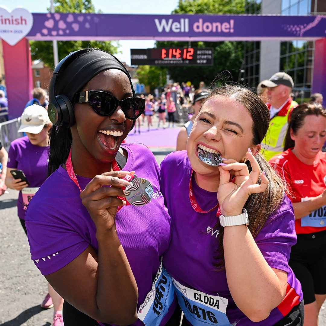 Two joyful women celebrate completing a race, wearing purple shirts, holding medals near their mouths with a finish line in view.